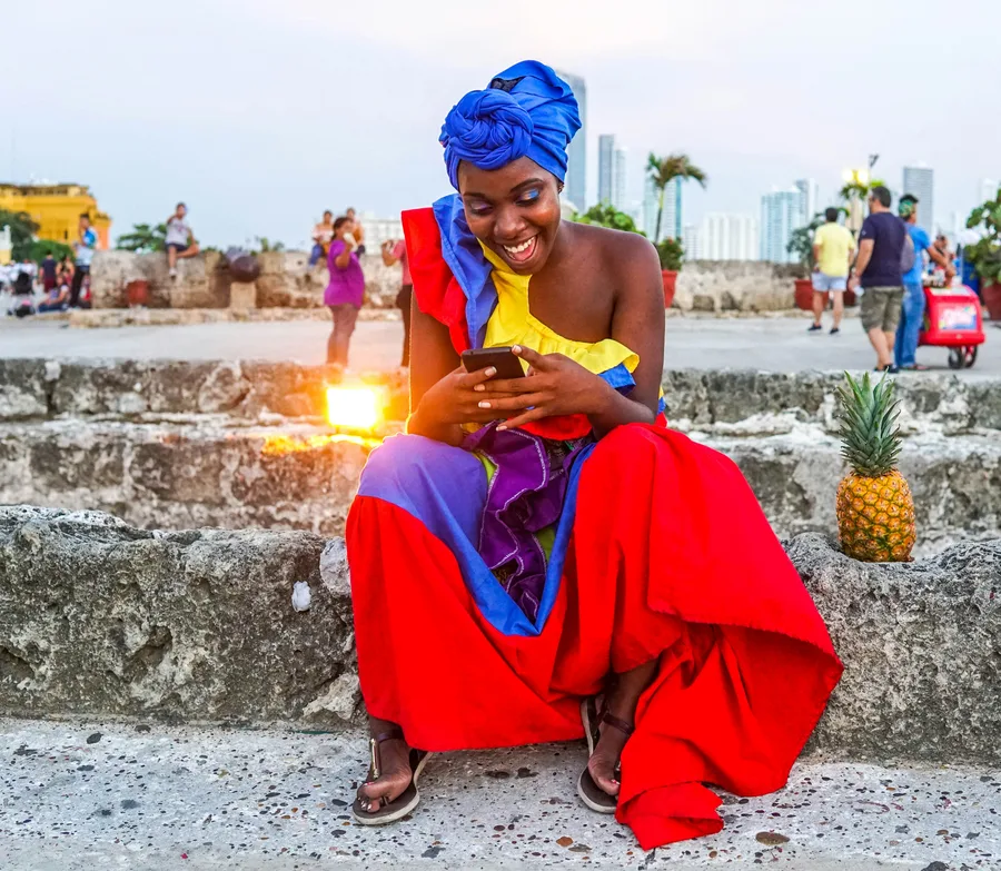 Palenquera con vestido tradicional de colores y móvil, sentada sobre las murallas de Cartagena al atardecer